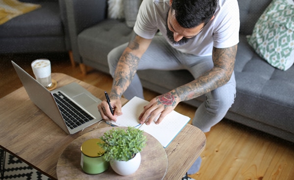 A man with tattoos writing in a notebook at a coffee table with a laptop and a cup of coffee, in a cozy living room, enjoying his Sunday habits.