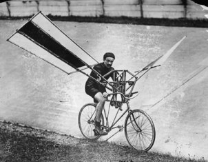 A man takes a risk riding a bicycle with a glider in this old photo.