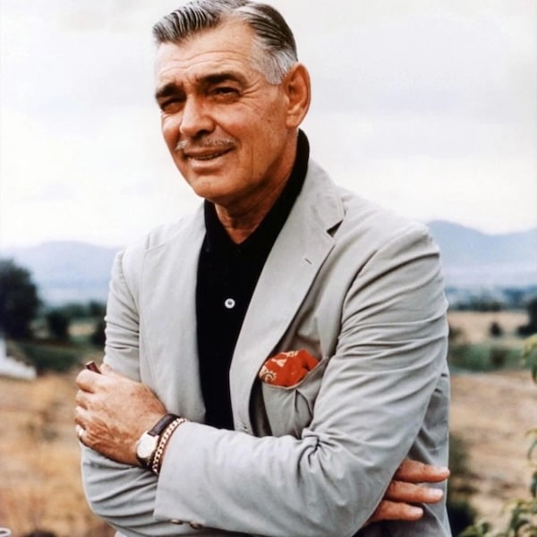 Clark Gable with gray hair and a mustache stands outdoors with arms crossed, showcasing men's fashion in a light gray suit jacket, black shirt, and red pocket square. A rural landscape and mountains are in the background.