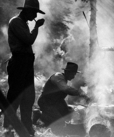 Black and white photograph of two men cooking with cast iron in the woods.