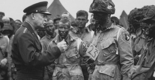 A military officer, reminiscent of Supreme Commander Eisenhower, speaks to a group of soldiers with camouflaged faces and uniforms, possibly preparing for a mission on D-Day during wartime.