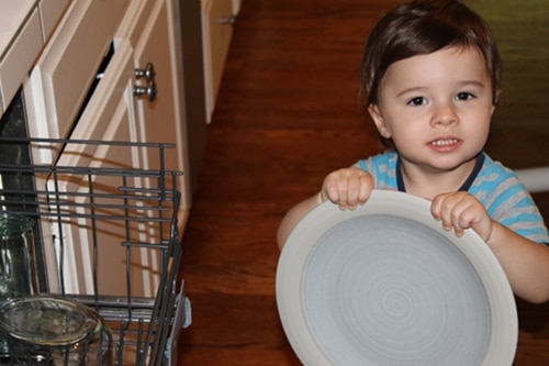 Young boy doing chores holding plate emptying dishwasher.