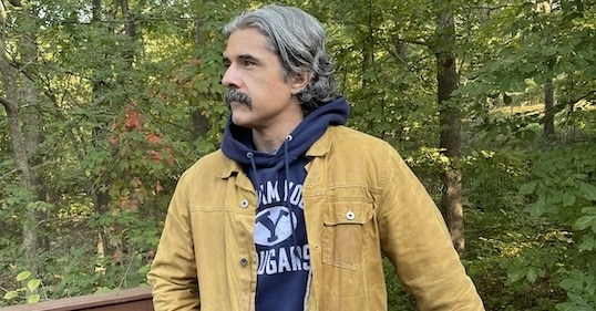 A man with gray hair and a mustache stands outdoors in front of trees, wearing a tan fall jacket over a blue hoodie.