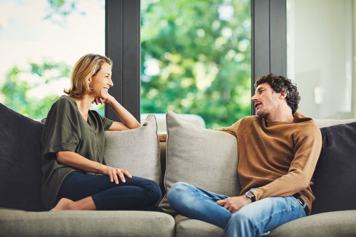 Two people sit on a sofa facing each other, engaged in conversation during a Marriage Checkup, with large windows and greenery visible in the background.