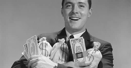 A smiling man in his 30s in a suit holds several bags marked with dollar signs and large paper bills, celebrating his personal finance goals.