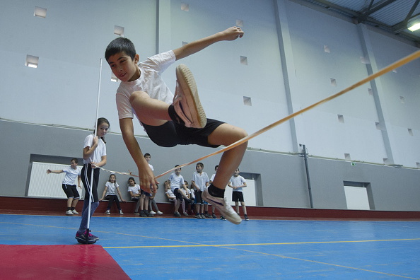 A boy is jumping over a high bar in a gym during P.E. class.